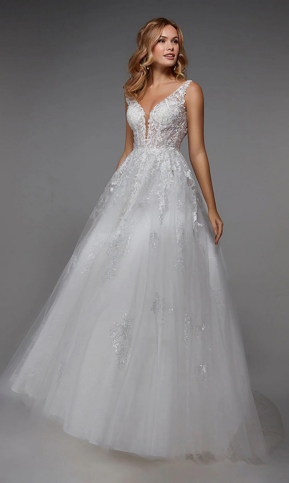 A woman wearing the Alyce 7039 long ivory dress—a sleeveless lace ball gown with a deep V-neckline and full tulle skirt—stands against a plain gray background.